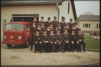 Eine Gruppe uniformierter Feuerwehrleute posiert für ein Foto vor einem Gebäude mit einem Feuerwehrwagen im Hintergrund.