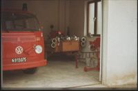 Ein alter roter Feuerwehrwagen steht in einer Garage mit weißen Wänden, begleitet von Feuerwehrausrüstung und Eimern.
