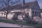 Ein altes Haus mit Schornstein und mehreren Fenstern steht in einem Hof, umgeben von kahlen Bäumen und einem Zaun. Das Haus scheint sich in einem schlechten Zustand zu befinden.