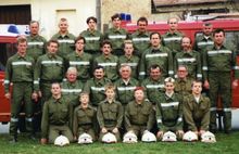 Eine Gruppe Feuerwehrleute in grünen Uniformen, einschließlich Männer und Frauen, posiert für ein Teamfoto mit einem roten Feuerwehrauto im Hintergrund.