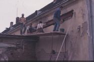 Vier Männer arbeiten an einem Gebäude. Sie stehen auf Gerüsten, zwei tragen Mörtel auf die Wand auf, einer trägt einen Eimer, und einer beobachtet.