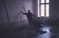Zwei Männer arbeiten in einem unfertigen Raum. Einer steht und hält ein Stück Holz, während der andere kniet und auf dem Boden arbeitet. Eine Ziegelmauer ist sichtbar.