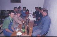 Eine Gruppe von Menschen sitzt um einen Tisch herum mit verschiedenen Flaschen und einem Glas, einige tragen Hüte. Ein Kind sitzt zwischen zwei Männern.