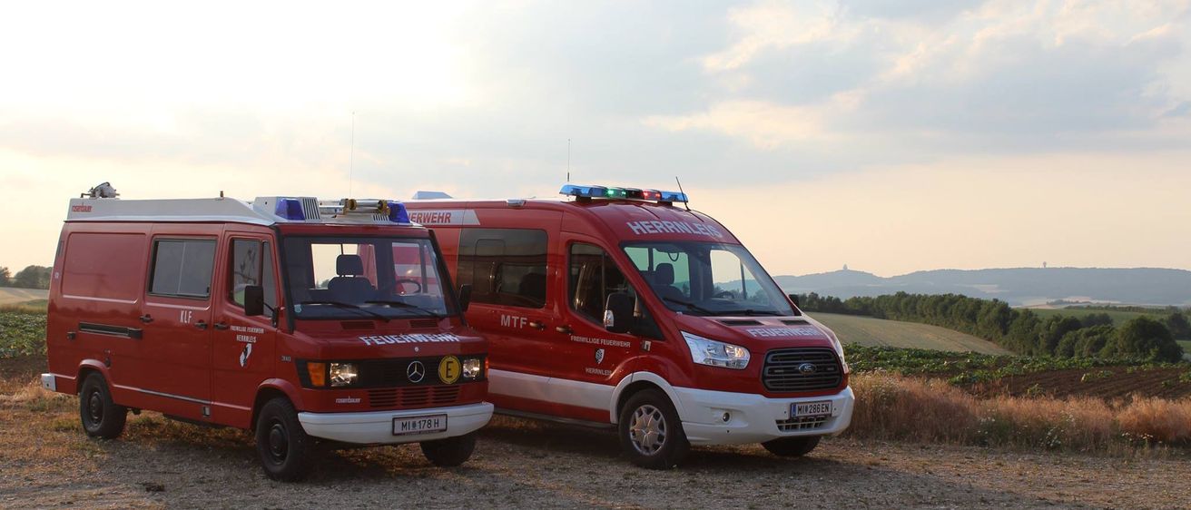 Zwei rote Krankenwagen sind auf einem Feldweg unter einem bewölkten Himmel geparkt. Einer ist mit 'Feuerwehr' und der andere mit 'Herrnwehr' beschriftet.