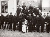 Eine Gruppe von Feuerwehrleuten in Uniformen posiert für ein Foto mit einer Frau vor einem Gebäude und einer Fahne.