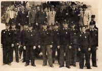 Ein altes Foto zeigt eine Gruppe uniformierter Männer, die in zwei Reihen stehen. Einige tragen Hüte und Krawatten, andere Armbinden. Sie scheinen in einer formellen Umgebung für ein Foto zu posieren.