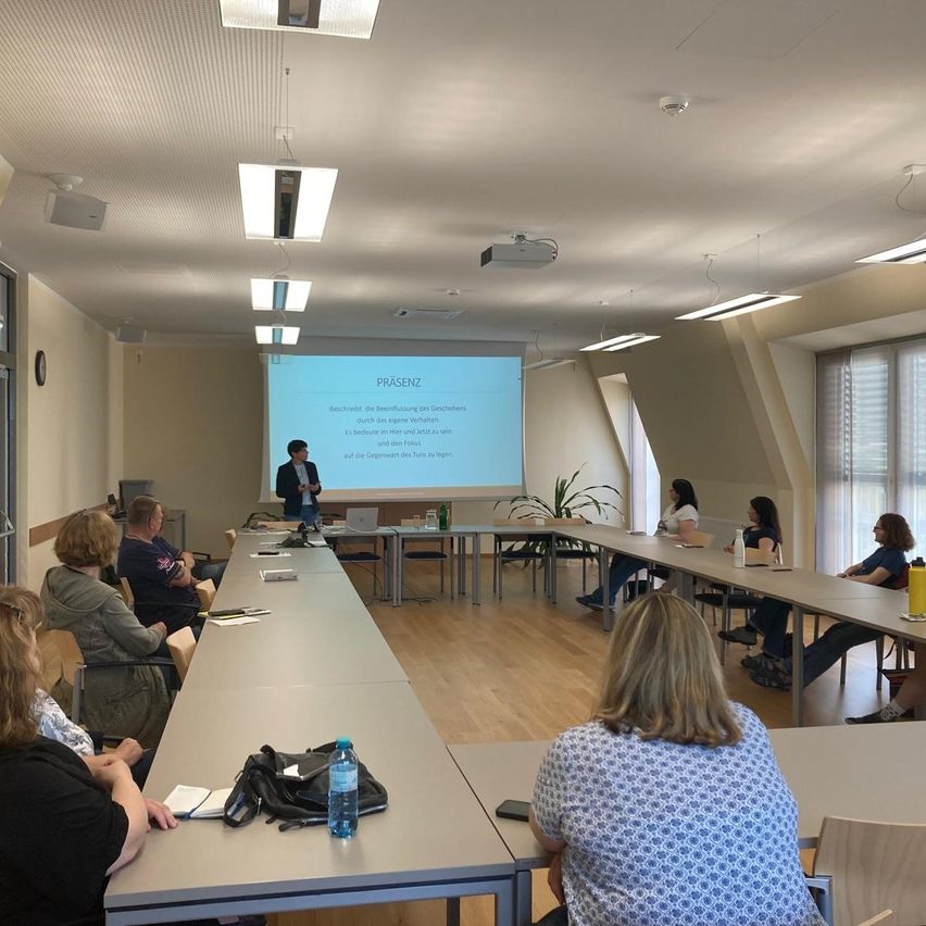 Ein Klassenzimmer mit einer Präsentationsleinwand, einem Mann, der präsentiert, und mehreren Frauen, die an Tischen sitzen und zuhören. Der Raum hat Holzböden, Deckenleuchten und einen Projektor.