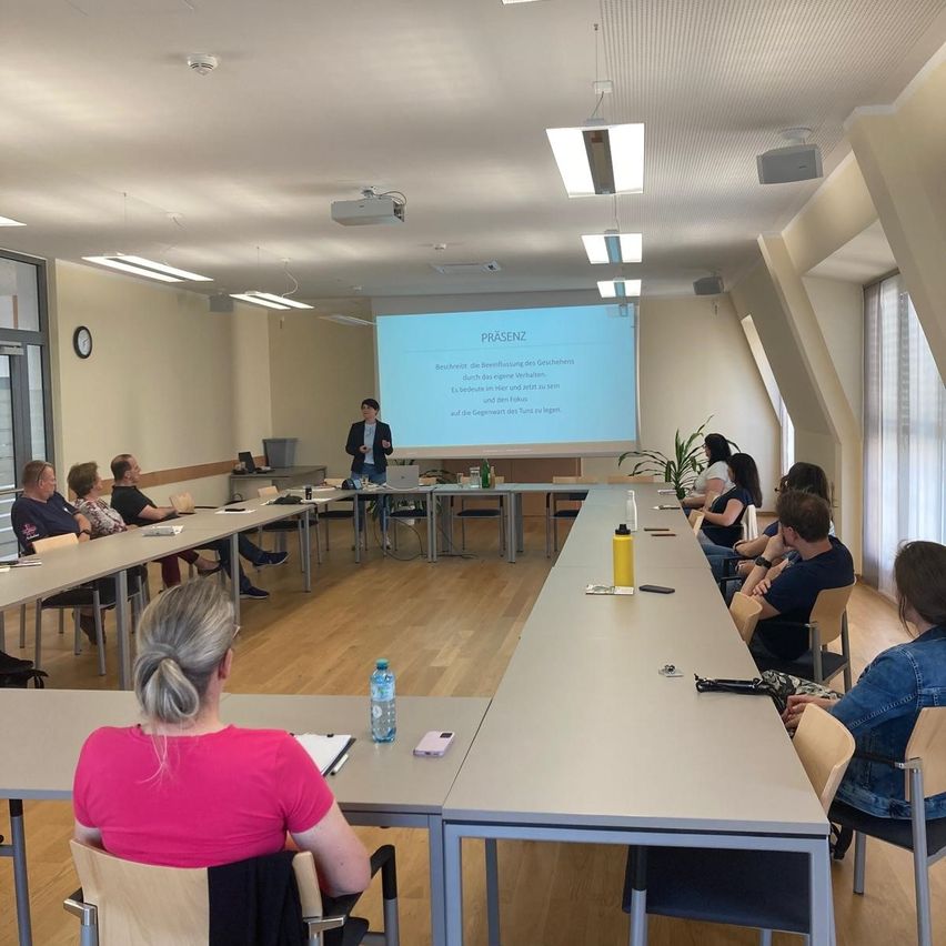 Ein Klassenzimmer mit einer Projektionsleinwand, auf der eine Präsentation mit dem Titel 'Präsenz' angezeigt wird. Eine Person steht an einem Tisch mit einem Laptop und spricht eine Zuhörerschaft an, die um Tische herumsitzt.