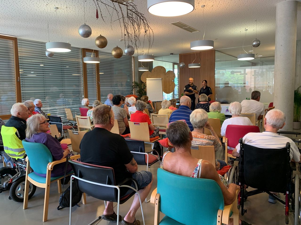 Eine Gruppe älterer Menschen sitzt in einem Raum auf Stühlen. Vor ihnen steht eine Tafel und zwei Polizeibeamte. Der Raum hat Deckenlampen und Glasfenster.