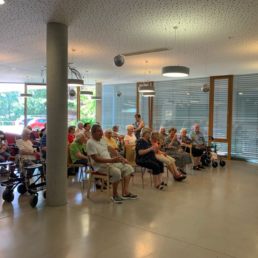 Eine Gruppe älterer Menschen sitzt in Stühlen in einer Halle mit Glasfenstern. Eine Frau macht ein Foto mit ihrem Handy.