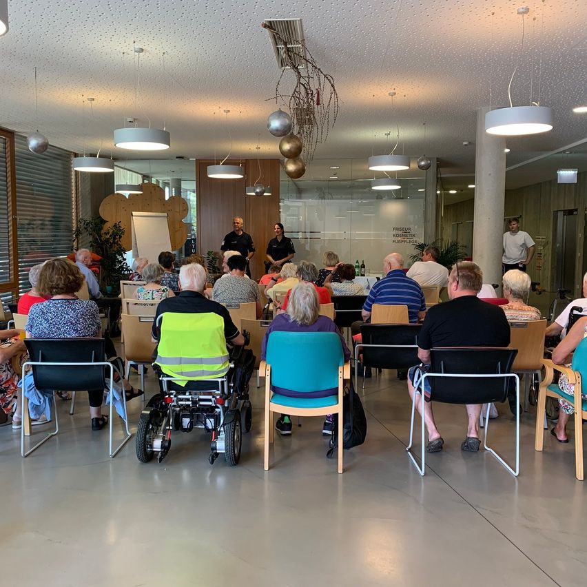 Eine Gruppe von Menschen sitzt in einem Konferenzraum auf Stühlen. Eine Person in einer Warnweste benutzt einen Rollstuhl. Zwei Personen stehen vor einer Tafel.