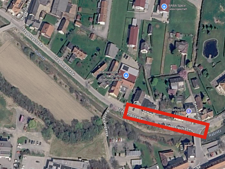 Luftaufnahme einer kleinen Stadt mit verschiedenen Gebäuden, Straßen und einem roten Rechteck, das einen bestimmten Bereich hervorhebt. Die Stadt umfasst Wohnhäuser, einen Blumenladen und einen Teich.