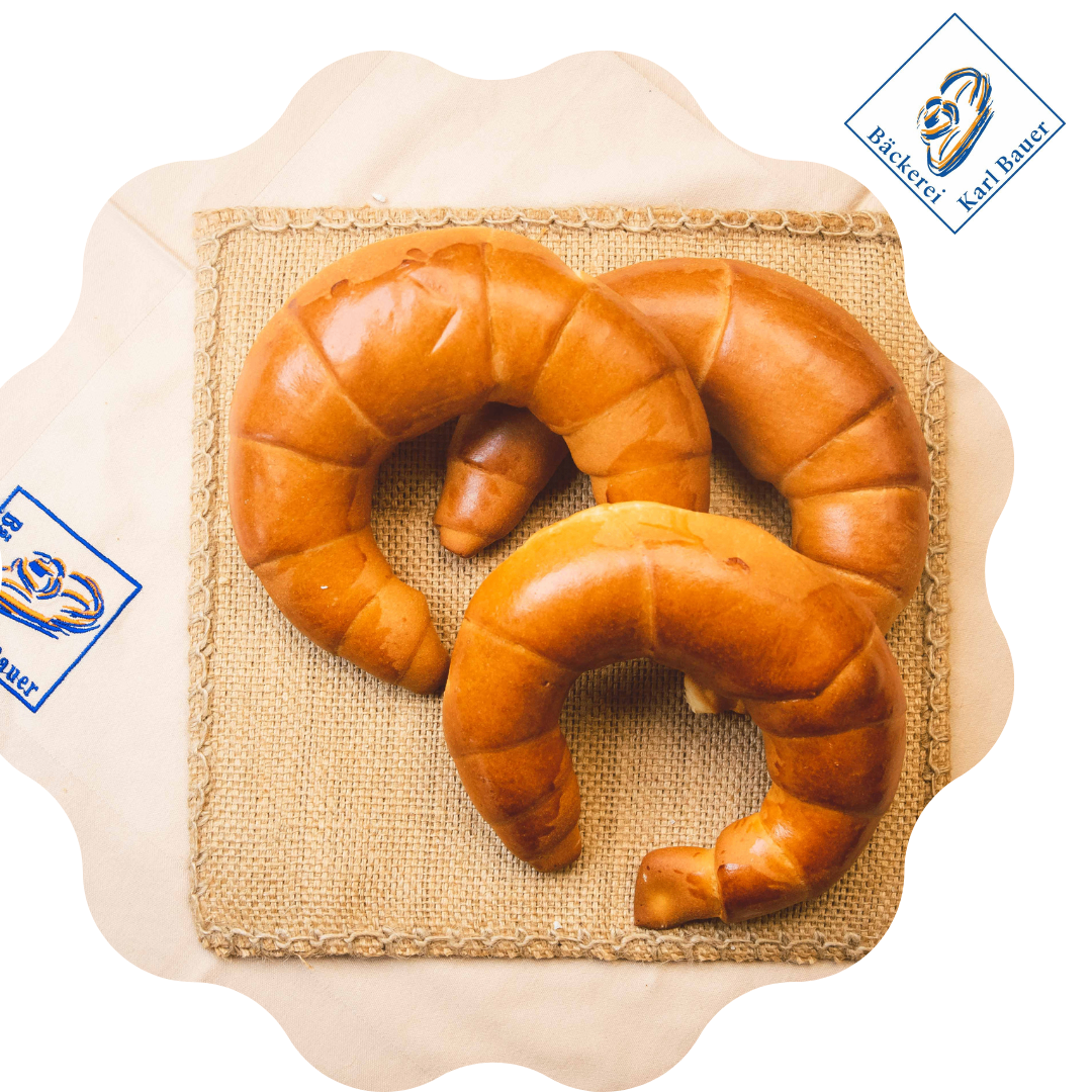 Zwei Croissants liegen auf einem Jutetuch auf einem beigefarbenen Umschlag. Die Croissants sind goldbraun und leicht gebogen. In der oberen rechten Ecke befindet sich ein Logo.