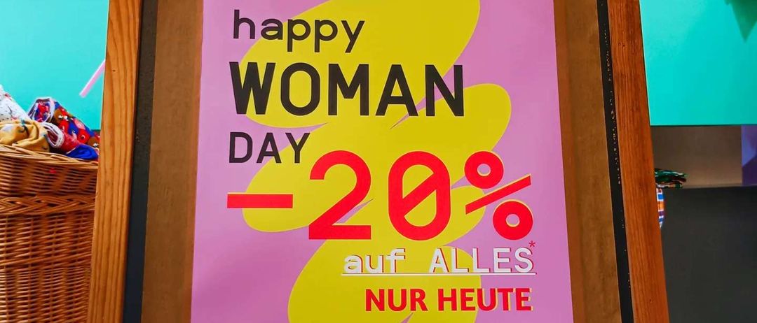 Ein rosafarbenes Plakat auf einem Holzrahmen kündigt einen 20%igen Rabatt für den Frauentag an, der nur heute gültig ist. Das Plakat zeigt Luftballons und einen Korb im Hintergrund.