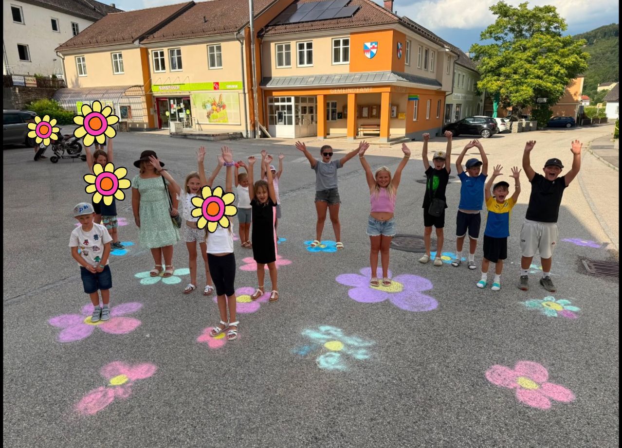 Eine Gruppe von Kindern und Erwachsenen steht in einem Kreis auf einer Straße mit auf dem Boden gezeichneten Blumen. Sie heben die Hände und lächeln. Hinter ihnen steht ein Gebäude mit einem Schild und einem geparkten Auto.