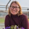 Eine Frau mit Brille lächelt, während sie einen Topf mit Blumen hält, in einem Gewächshaus stehend.