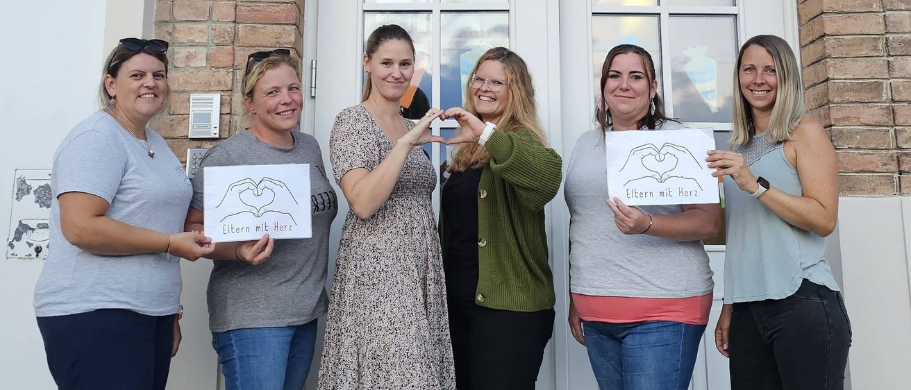 Fünf Frauen stehen vor einem Gebäude mit Schildern. Sie lächeln und posieren für ein Foto. Das Schild am Gebäude steht 'Hauptstraße 5'.