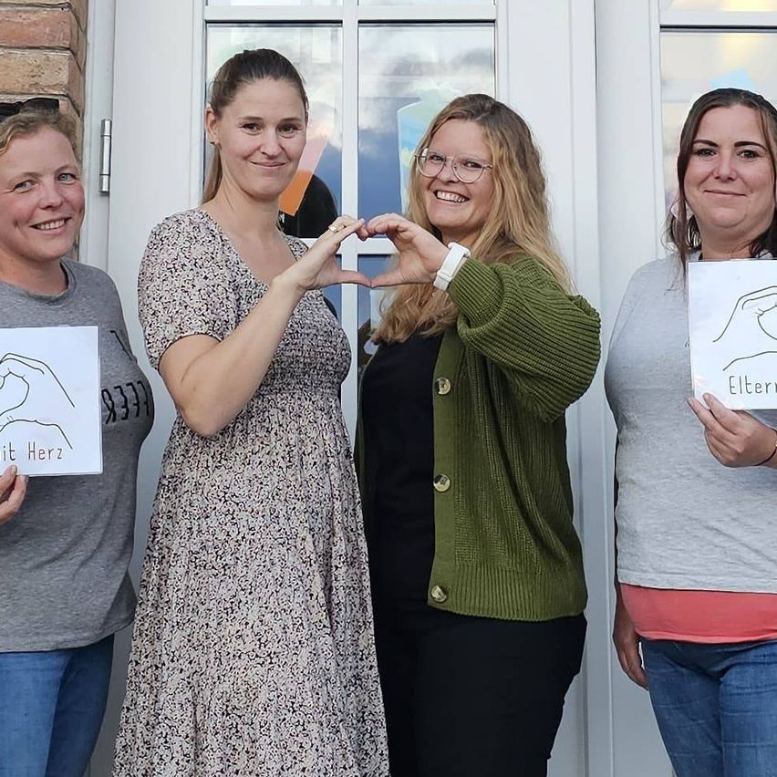 Fünf Frauen stehen vor einem Gebäude mit Schildern. Sie lächeln und posieren für ein Foto. Das Schild am Gebäude steht 'Hauptstraße 5'.