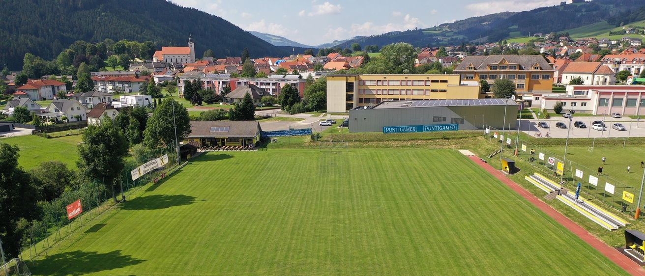 Ein großes Sportfeld mit einem Fußballtor, umgeben von Gebäuden, Bäumen und Bergen.