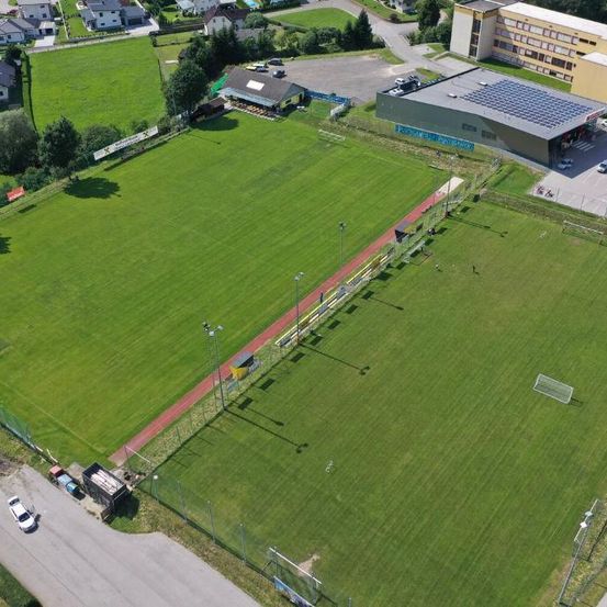 Ein Fußballfeld mit Laufbahn, Bänken und Straßenlaternen. Gebäude und Autos in der Umgebung.