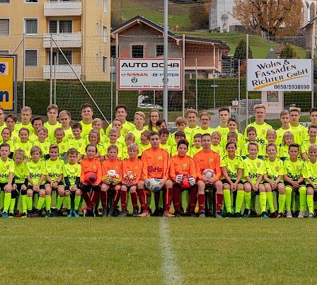 Eine Fußballmannschaft in neongrünen Trikots posiert für ein Gruppenfoto auf einem Feld. Gebäude und Bäume im Hintergrund.