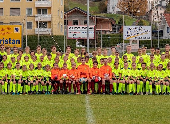 Eine Fußballmannschaft in neongrünen Trikots posiert für ein Gruppenfoto auf einem Feld. Gebäude und Bäume im Hintergrund.