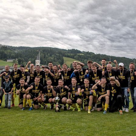 Eine Fußballmannschaft feiert auf einem Feld mit Bergen im Hintergrund. Sie halten einen Pokal und tragen Medaillen.