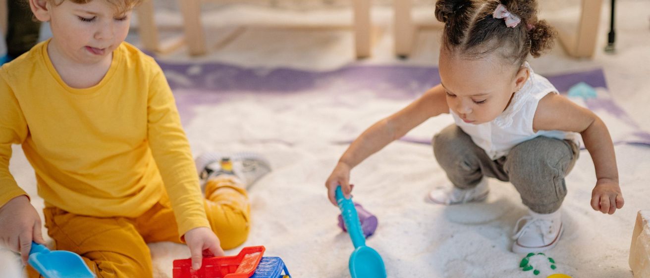 Zwei kleine Kinder spielen mit Spielzeug in einem Sandkasten, eines hält einen Spielzeuglastwagen und das andere eine Schaufel. Das Mädchen trägt ein weißes Kleid und der Junge ein gelbes Langarmshirt. Stühle sind im Hintergrund.