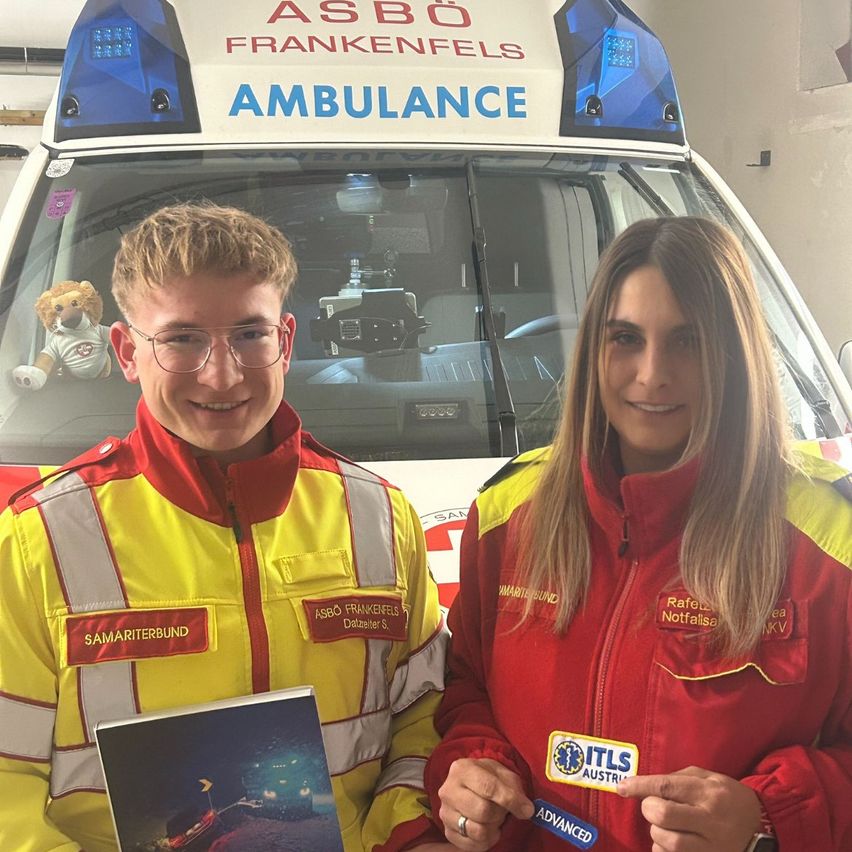Two individuals dressed in reflective emergency responder uniforms stand in front of an ambulance. The man holds a book and smiles while the woman holds a badge.