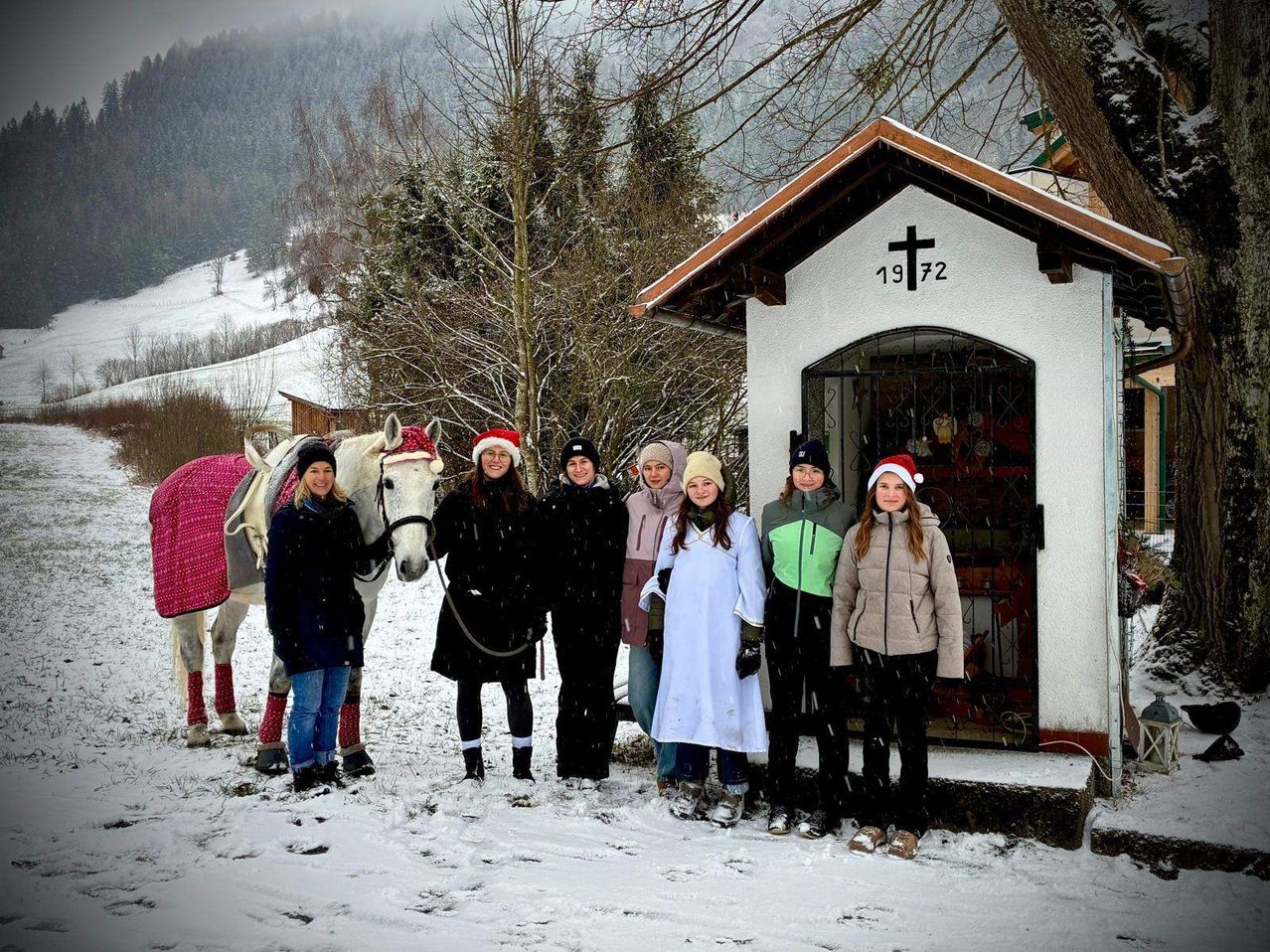 Eine Gruppe von Menschen in warmen Kleidern steht vor einer kleinen Kapelle mit einem Kreuz, im Schnee, mit einem Pferd in der Nähe.