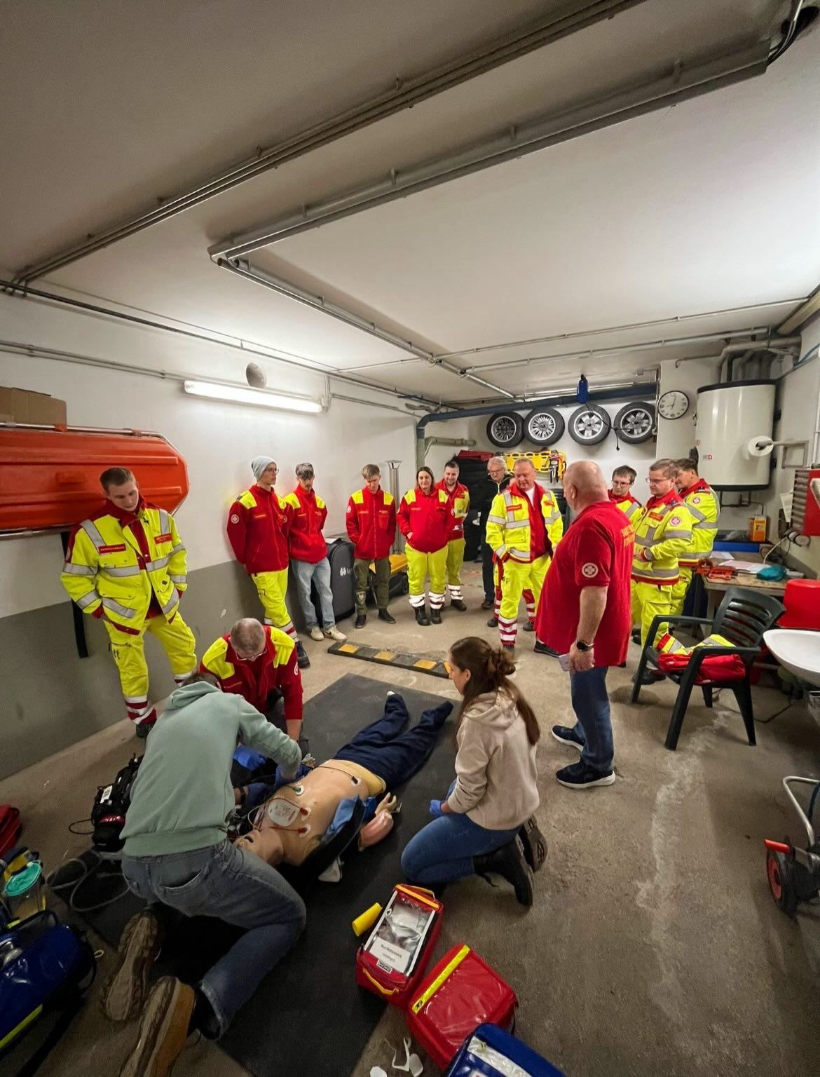 Eine Gruppe von Menschen in roten und gelben Uniformen übt Erste Hilfe in einem Raum mit einemDummy auf dem Boden. Einige stehen, andere knien. Eine Frau beobachtet.