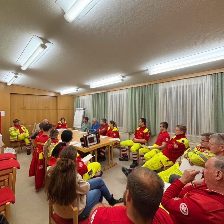 Eine Gruppe von Feuerwehrleuten in roten und gelben Uniformen sitzt um einen Tisch herum, möglicherweise in einer Besprechung. Der Raum hat weiße Wände, Holzschränke und grüne Vorhänge.