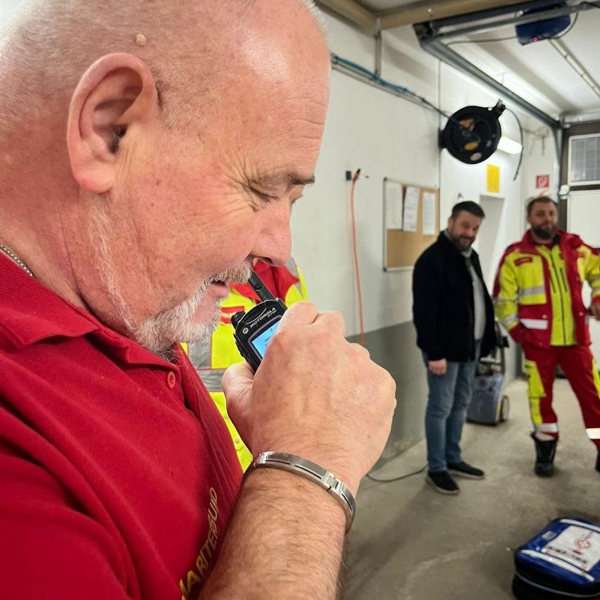 Ein Mann in einem roten Shirt hält ein Motorola-Walkie-Talkie und schaut nach unten. Zwei Männer in Sicherheitsanzügen stehen hinter ihm in einem Raum mit weißen Wänden.