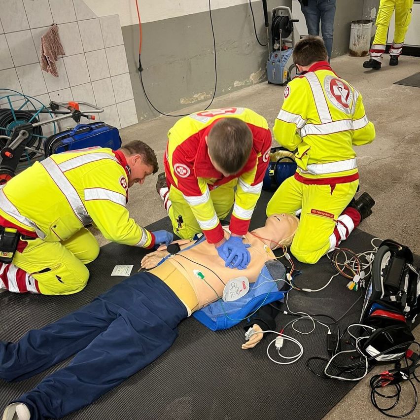 Drei Notfallsanitäter in gelben Uniformen kümmern sich um einen auf einer Matte liegenden Mannequin, der eine medizinische Notlage simuliert. Ausrüstung und Kabel sind umher verstreut.