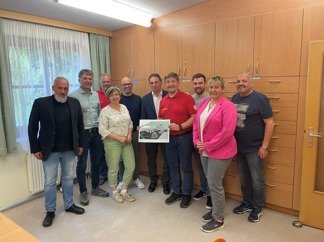 A group of people stand in a room with wooden cabinets. They hold a piece of paper showing a building plan.