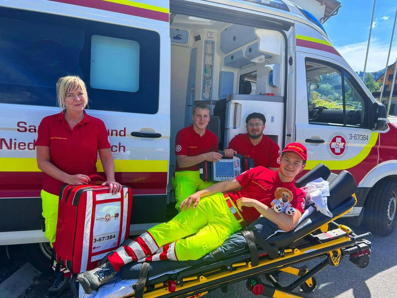 Vier Personen, darunter zwei Männer, stehen neben einem Krankenwagen. Ein Mann liegt auf einer Trage, und eine Frau hält eine rote Tasche. Ein anderer Mann hält ein Gerät.