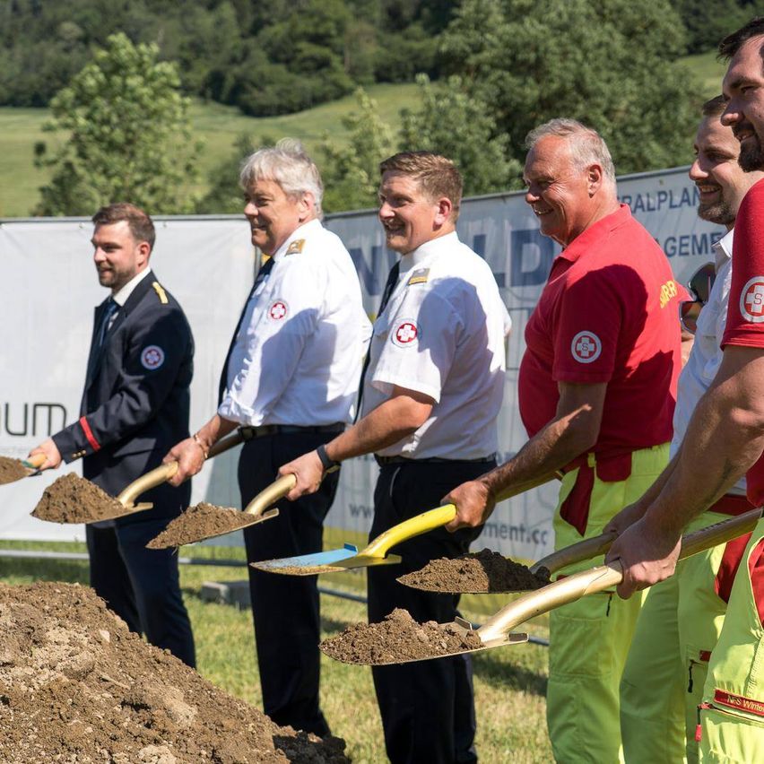 Eine Gruppe von Männern in Uniformen und Schutzausrüstung, die Schaufeln halten, stehen um einen Haufen Erde herum. Hinter ihnen ein Banner und Bäume. Sie scheinen sich auf einer Grundsteinlegungszeremonie zu befinden.