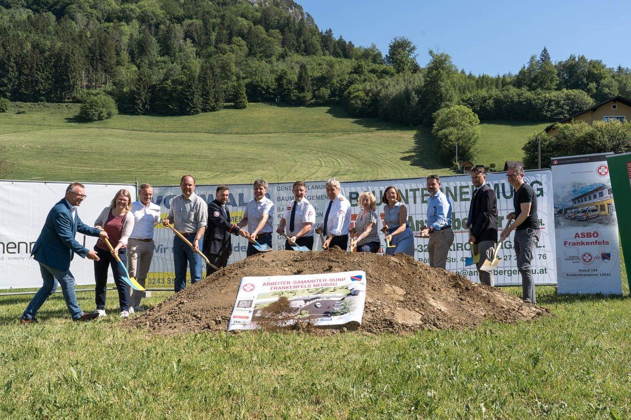 Eine Gruppe von Menschen steht um einen Haufen Erde herum und hält Schaufeln. Dahinter steht ein Banner mit den Aufschriften "Baumpflege" und "Generative Bau". Sie lächeln und schauen in die Kamera.