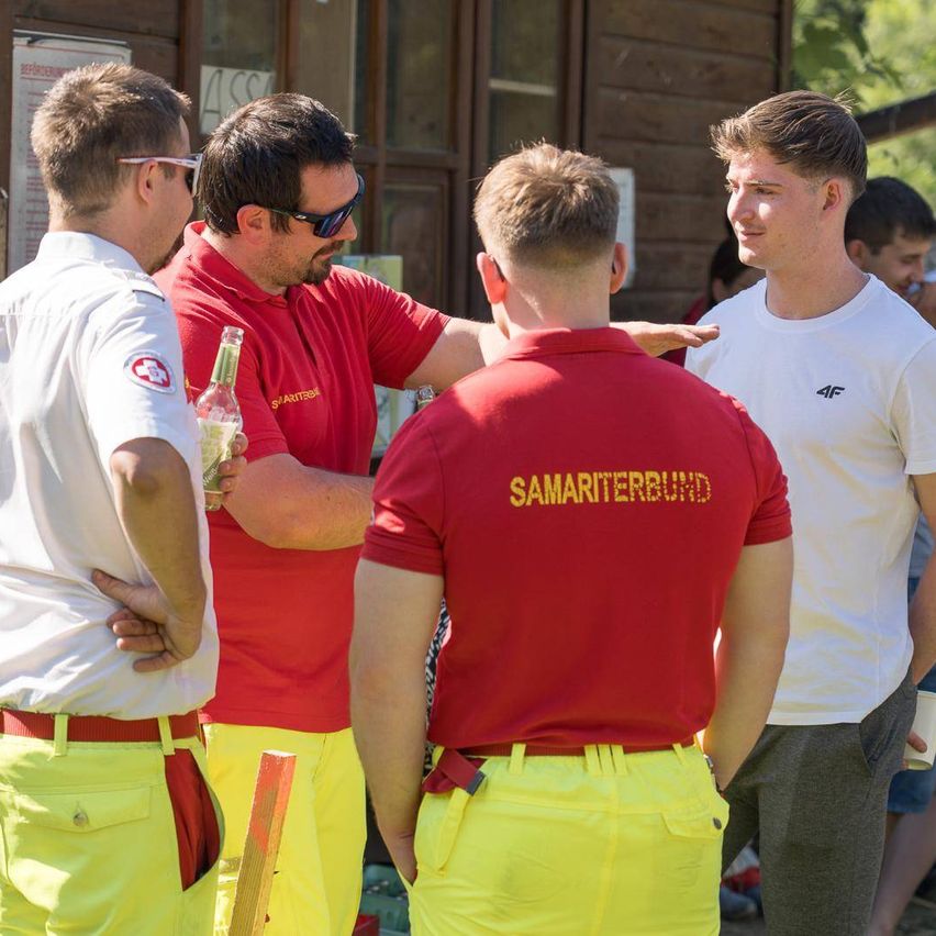 Fünf Männer stehen in einer Reihe, zwei tragen rote und gelbe Uniformen, einer hält eine Flasche. Die beiden vorne tragen weiße Hemden. Die roten Hemden sagen 'Samariterbund'.