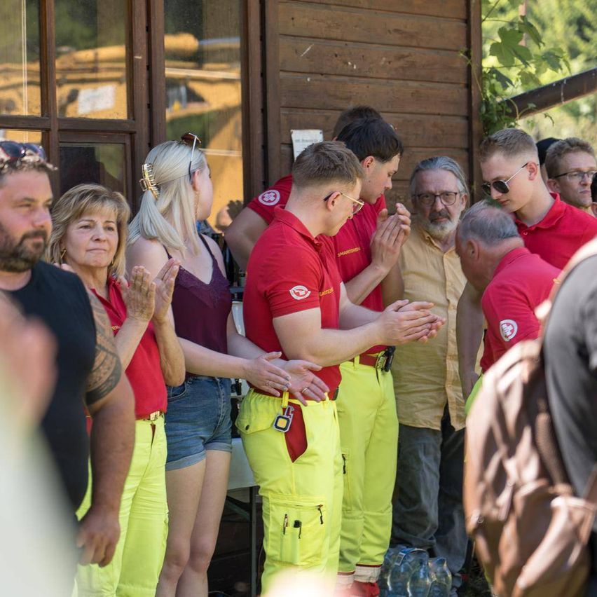 Eine Gruppe von Menschen in roten und gelben Uniformen, möglicherweise Feuerwehrleute, stehen und klatschen. Sie sind vor einem Gebäude. In der Mitte steht eine Frau mit einem lilafarbenen ärmellosen Oberteil.
