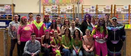 Eine Gruppe von Menschen in 80er- und 90er-Jahre-Kostümen posiert für ein Foto in einem mit Luftballons und Wimpelketten geschmückten Raum.