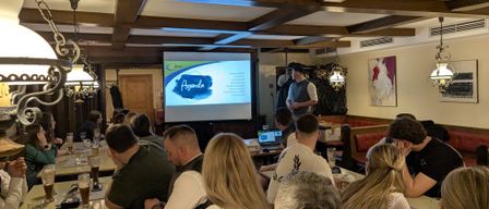 In einem Raum mit Holzdecken präsentiert ein Mann eine Folie auf einem Projektor. Eine Menschenmenge sitzt vor ihm, einige trinken Bier.