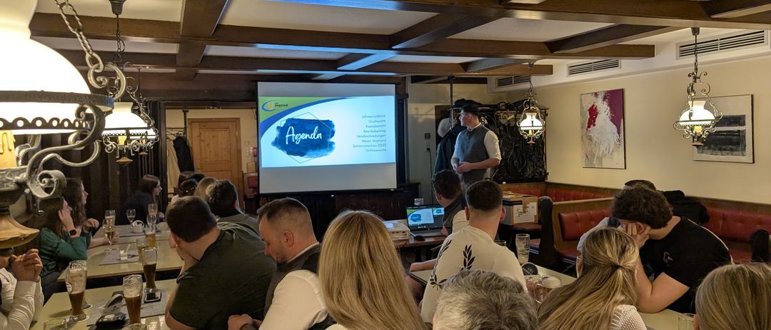In einem Raum mit Holzdecken präsentiert ein Mann eine Folie auf einem Projektor. Eine Menschenmenge sitzt vor ihm, einige trinken Bier.