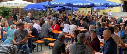 Eine überfüllte Biergarten im Freien mit blauen Sonnenschirmen und Tischen. Viele Menschen sitzen und trinken. Werbebanner hängen über ihnen.
