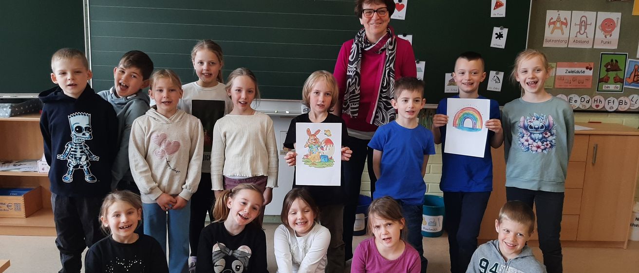 Eine Lehrerin steht mit einer Gruppe von Kindern, die Zeichnungen halten, in einem Klassenzimmer. Dahinter steht eine Tafel mit den Worten 'Dienstag, 8. April' und einem Datum.