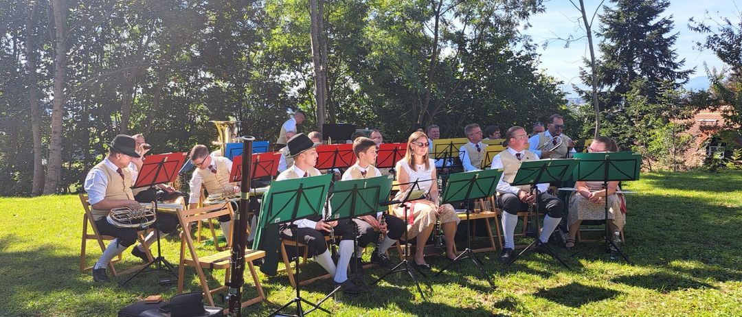 Eine Gruppe von Musikern in traditioneller Kleidung sitzt auf Stühlen in einem Grasbereich, möglicherweise bei einer Freiluftaufführung. Die Musiker haben verschiedene Instrumente, darunter Trompeten und Tuben.