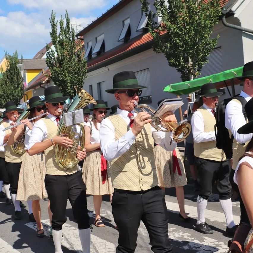 Eine Blaskapelle in traditioneller Kleidung spielt Instrumente auf einer Straße, mit Gebäuden und Bäumen im Hintergrund.