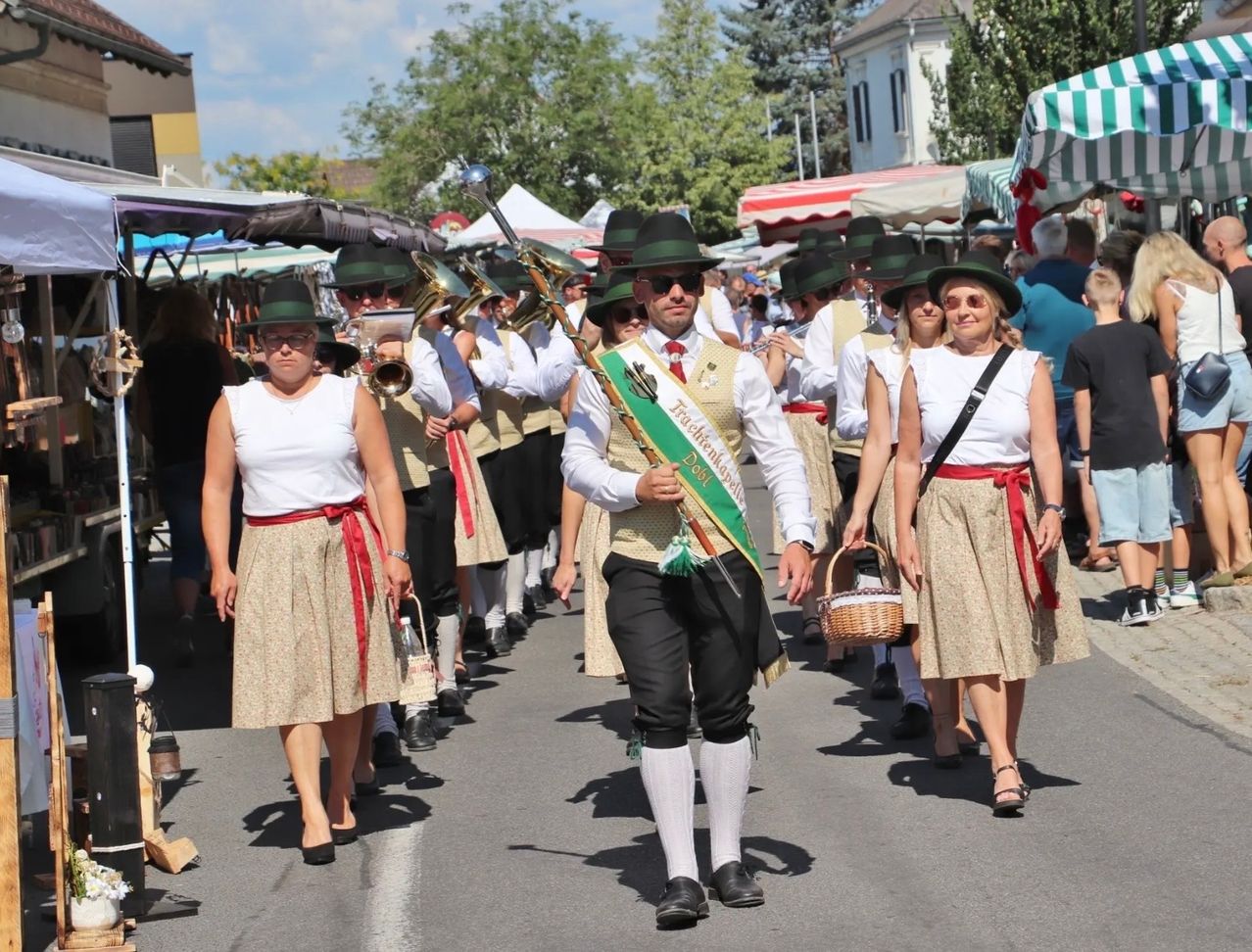 Eine Gruppe von Menschen in traditioneller Kleidung marschiert bei einem Umzug, begleitet von Musikern, die Blechblasinstrumente spielen. Die Szene spielt sich draußen an einem sonnigen Tag ab.