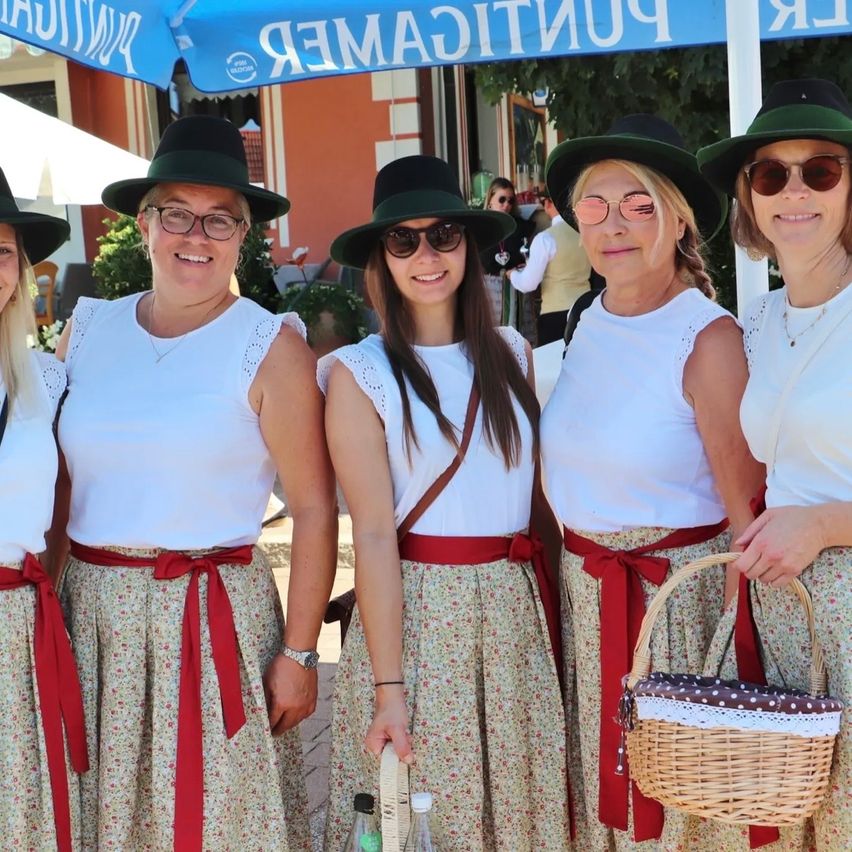 Fünf Frauen in passenden Outfits mit roten Bändern um die Taille stehen zusammen unter einem blauen Schild. Sie lächeln und tragen Hüte und Sonnenbrillen.