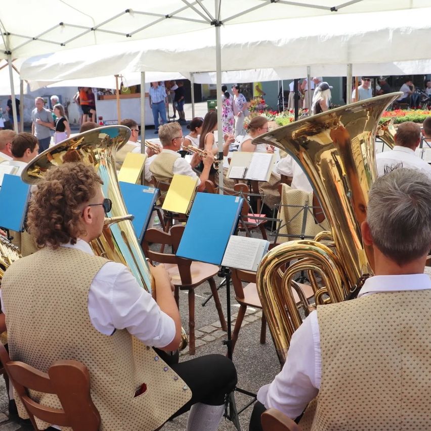 Ein Brass-Band spielt in einer Freiluft-Veranstaltung mit einem weißen Zelt im Hintergrund. Die Musiker spielen verschiedene Instrumente, darunter Tubas und Trompeten.